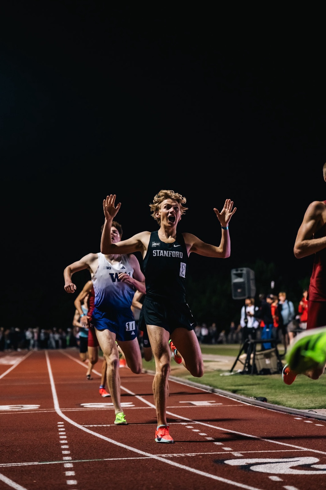 Zane crossing the finish line at night in a Stanford singlet, arms raised, ahead of a Washington runner